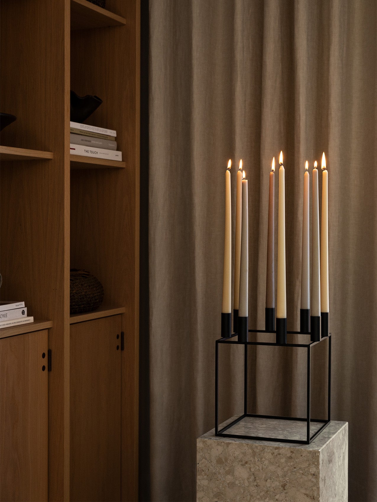 The black metal Kubus 8 candle holder by Lassen, holding eight lit candles, sits on a stone plinth in a stylish interior.
