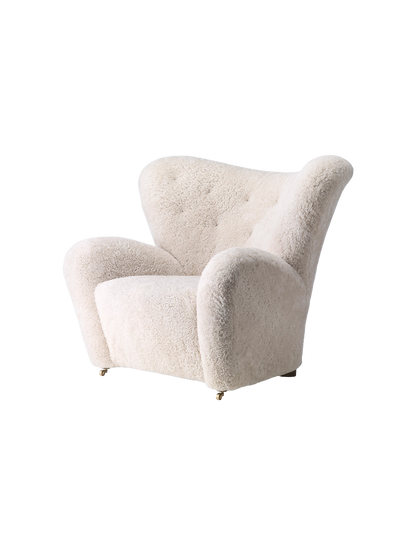 Side view of The Tired Man Lounge Chair in off-white sheepskin, showing its curved high back, large arms, and brass wheels.