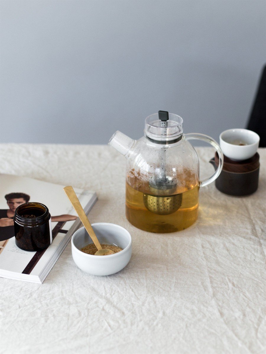 The glass Kettle Teapot by Norm Architects steeping tea on a linen tablecloth, next to a teacup, magazine and sugar bowl.