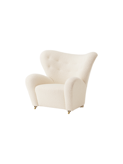 An angled view of The Tired Man Lounge Chair in a textured off-white textile, showing its curved high back and brass castors.