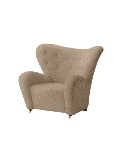 Angled view of The Tired Man Lounge Chair in light brown textile, featuring high curved back, plush arms, and brass castors