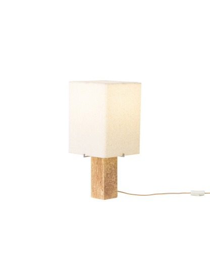Angled view of the lit Nonna Table Lamp, featuring a square travertine base and a warm, glowing sand-coloured linen shade.