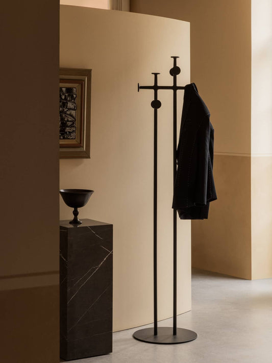 A black Afteroom Coat Stand with a jacket, styled in a hallway next to a tall black marble Plinth with a Pavot Bowl.