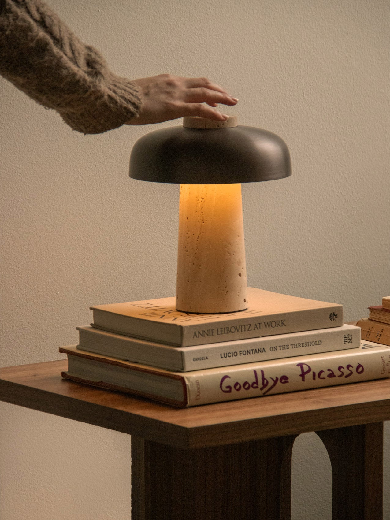 A hand adjusts the lit Reverse Table Lamp, Portable, which sits on a stack of art books on a wooden side table.
