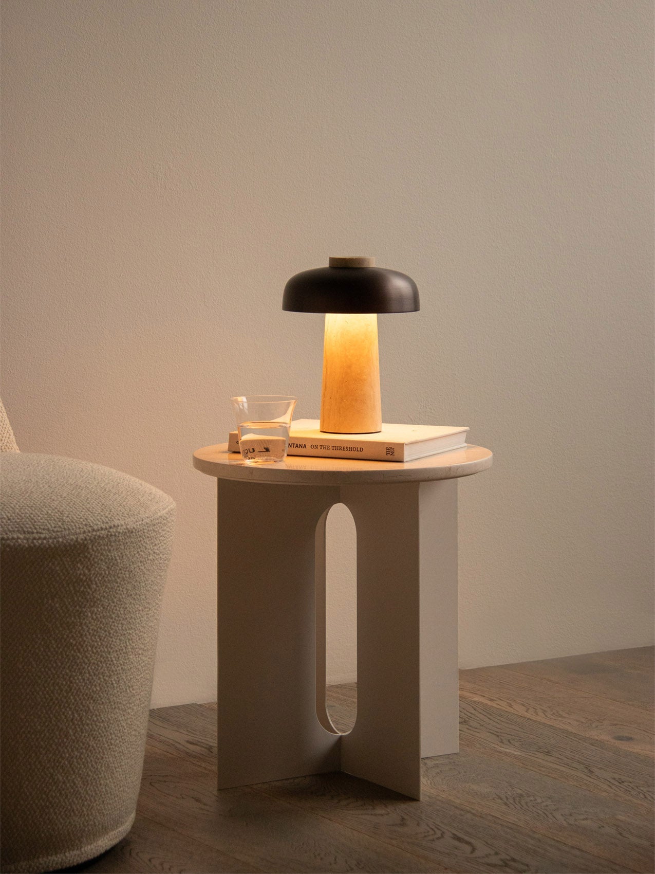 The lit Reverse Table Lamp, Portable sits on an Androgyne Side Table with a book and glass, beside a textured armchair.