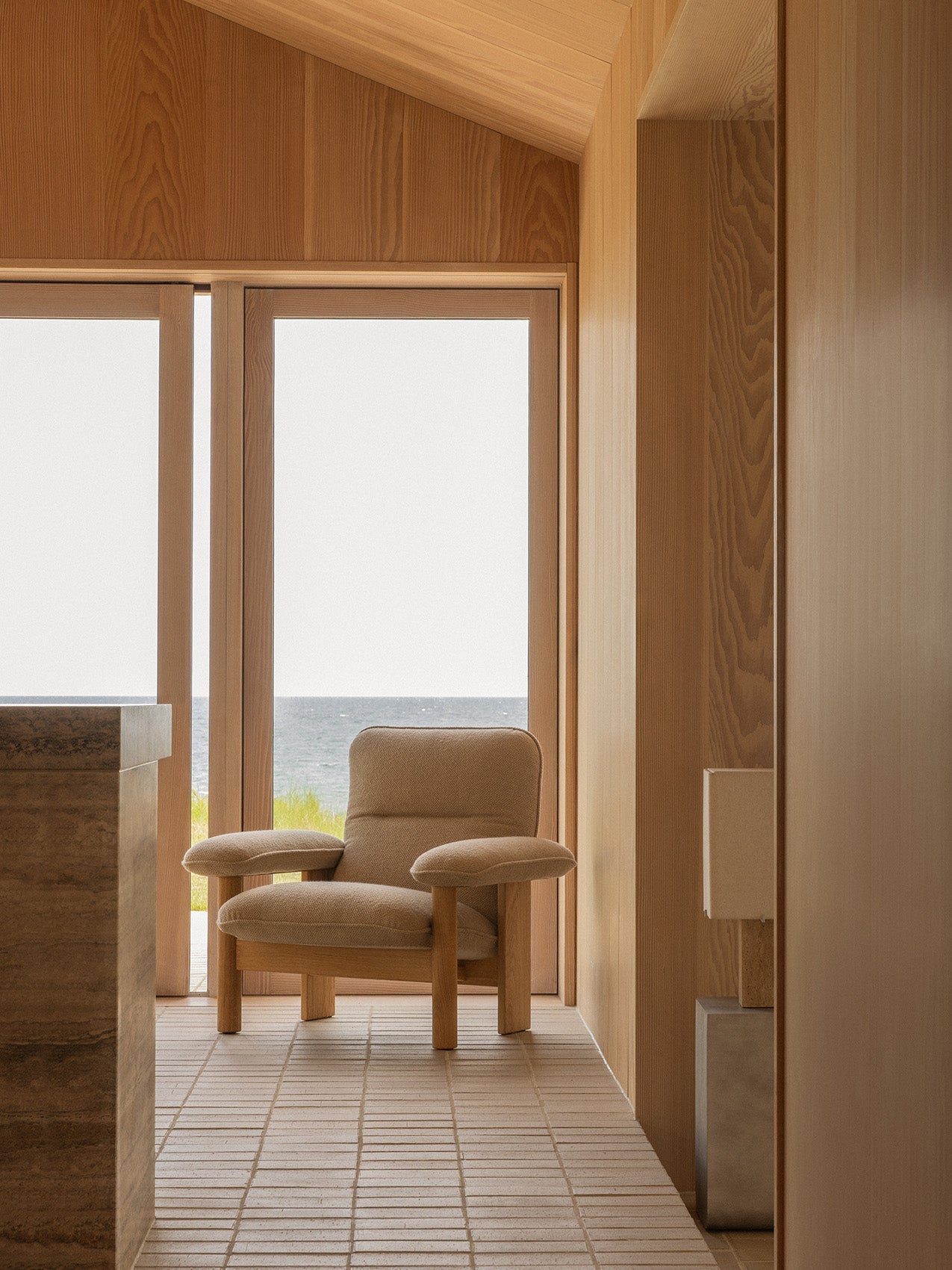 A beige Brasilia Lounge Chair with a light wood frame sits in a wood-panelled room, looking out of a window onto the sea.