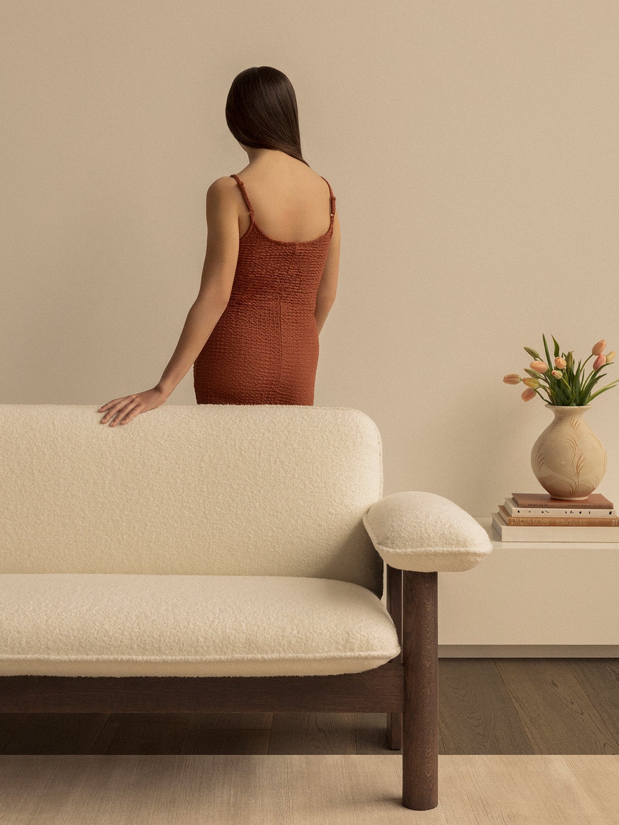 Back view of a woman resting her hand on a cream Brasilia Sofa. To the right, a vase of tulips sits on a stack of books.