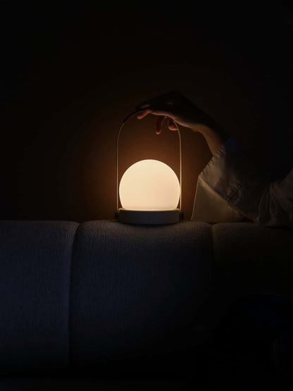 The Carrie Portable Lamp with opal glass shade and leather handle glows warmly in a dark room as a hand reaches for it.
