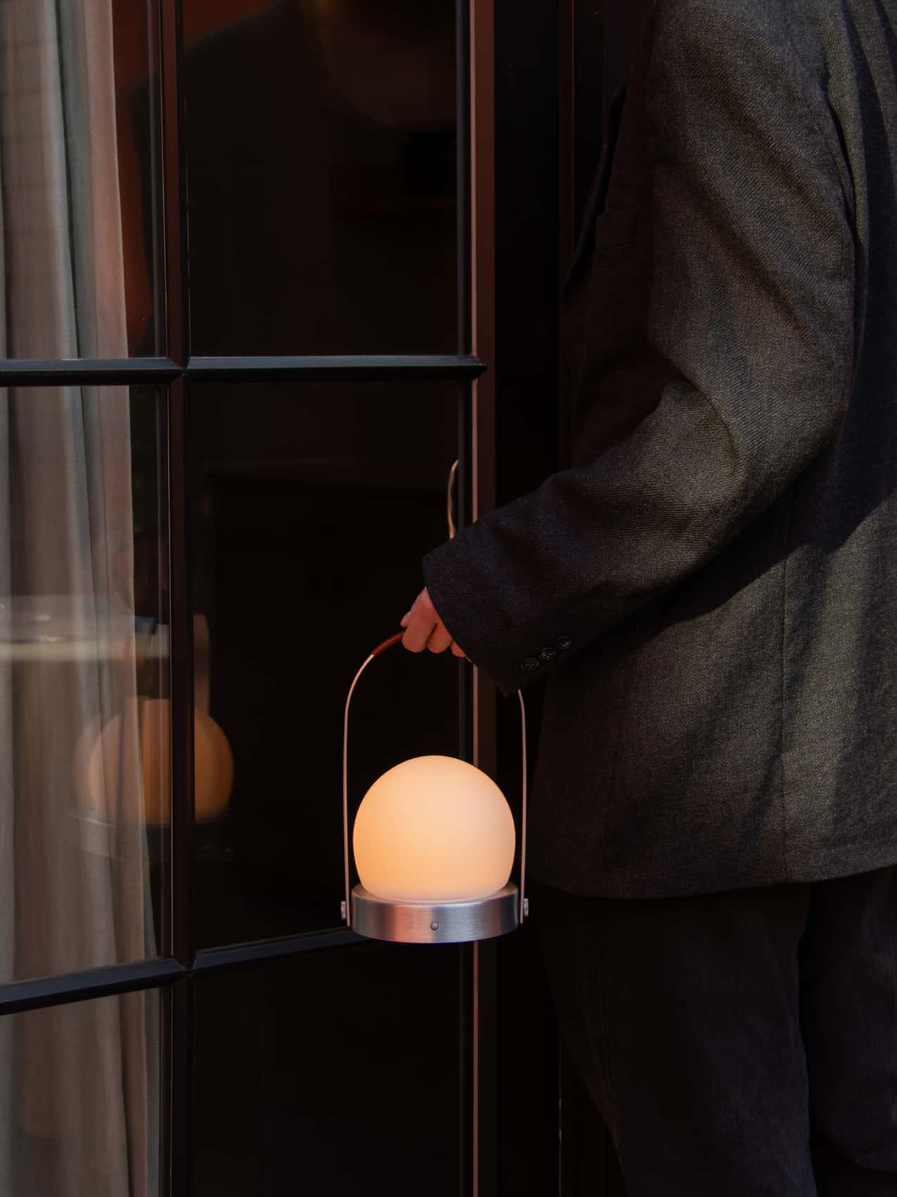 A glowing Carrie Portable Lamp with a brushed aluminium base and leather handle, carried by a person in a dark jacket.