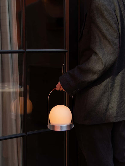 A glowing Carrie Portable Lamp with a brushed aluminium base and leather handle, carried by a person in a dark jacket.