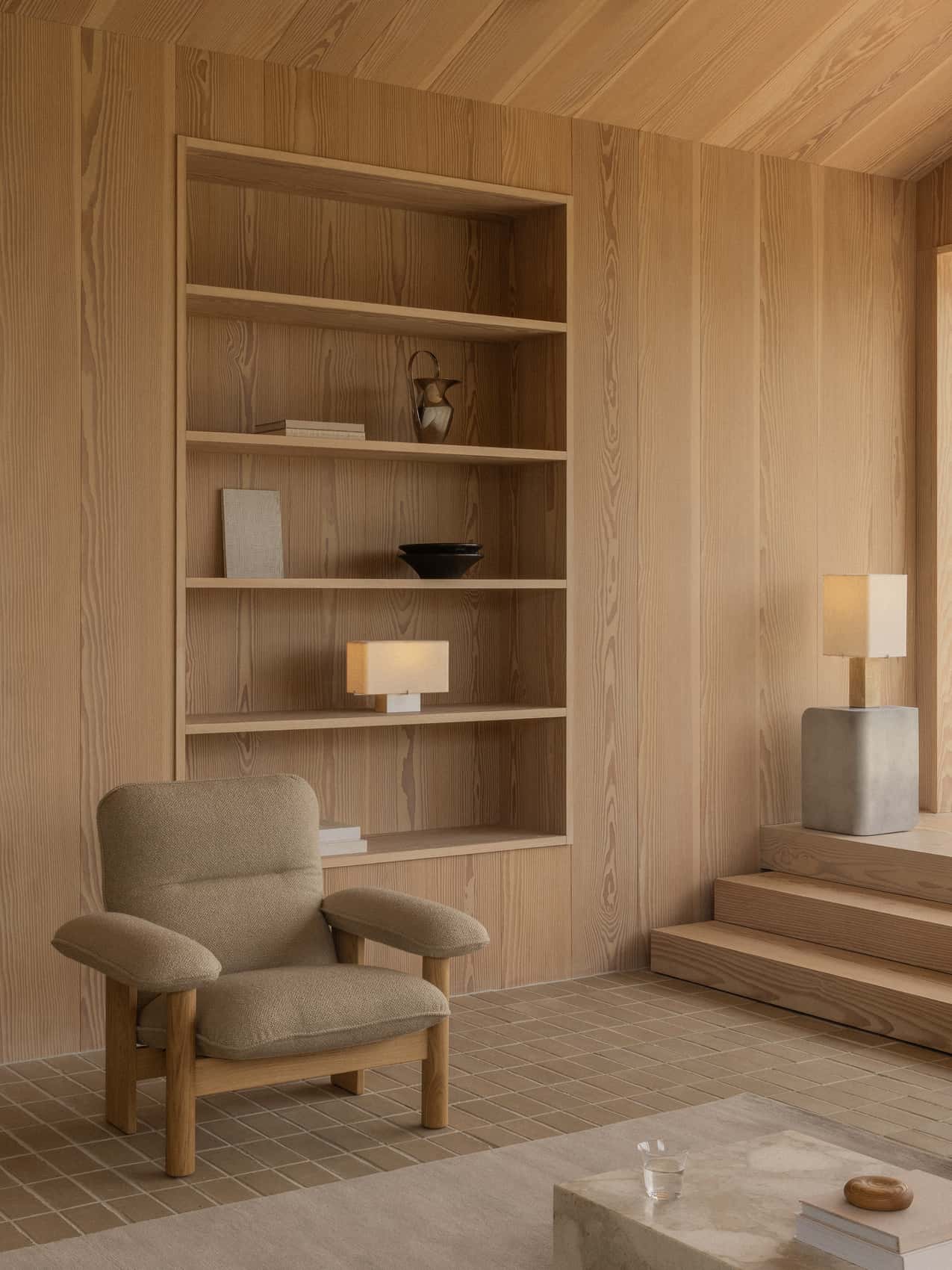 A wood-panelled room featuring a beige Brasilia Lounge Chair and built-in shelves, with a lit Nonna Table Lamp on display.