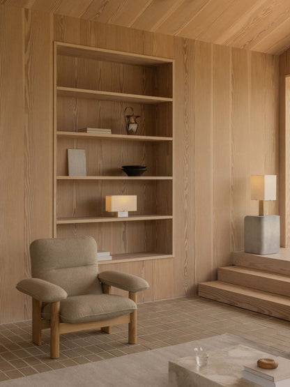 A wood-panelled room featuring a beige Brasilia Lounge Chair and built-in shelves, with a lit Nonna Table Lamp on display.