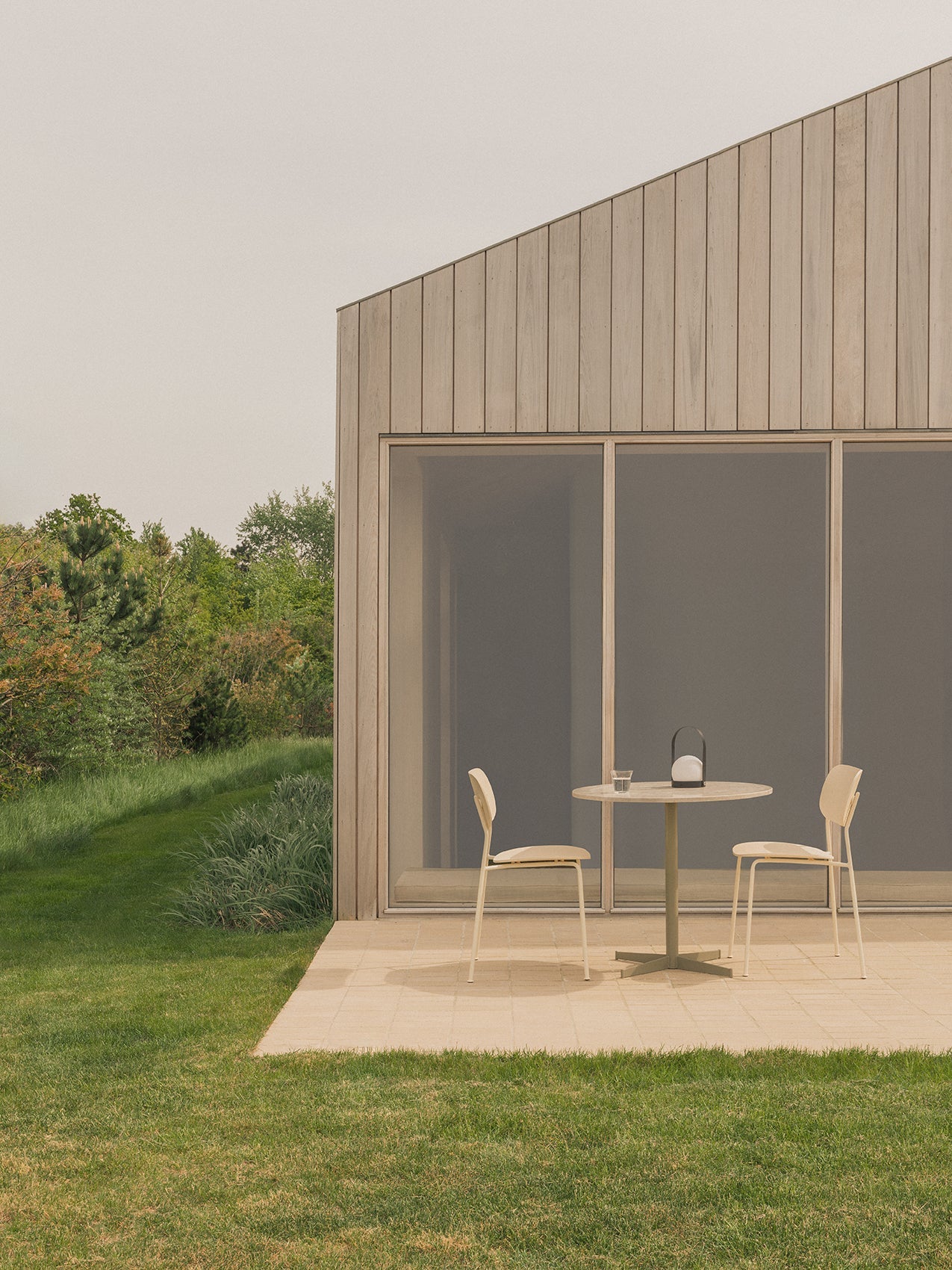 Outdoor patio with a Nook Dining Table, two Co Dining Chairs, and a Carrie Lamp, set against a modern wood-panelled building.