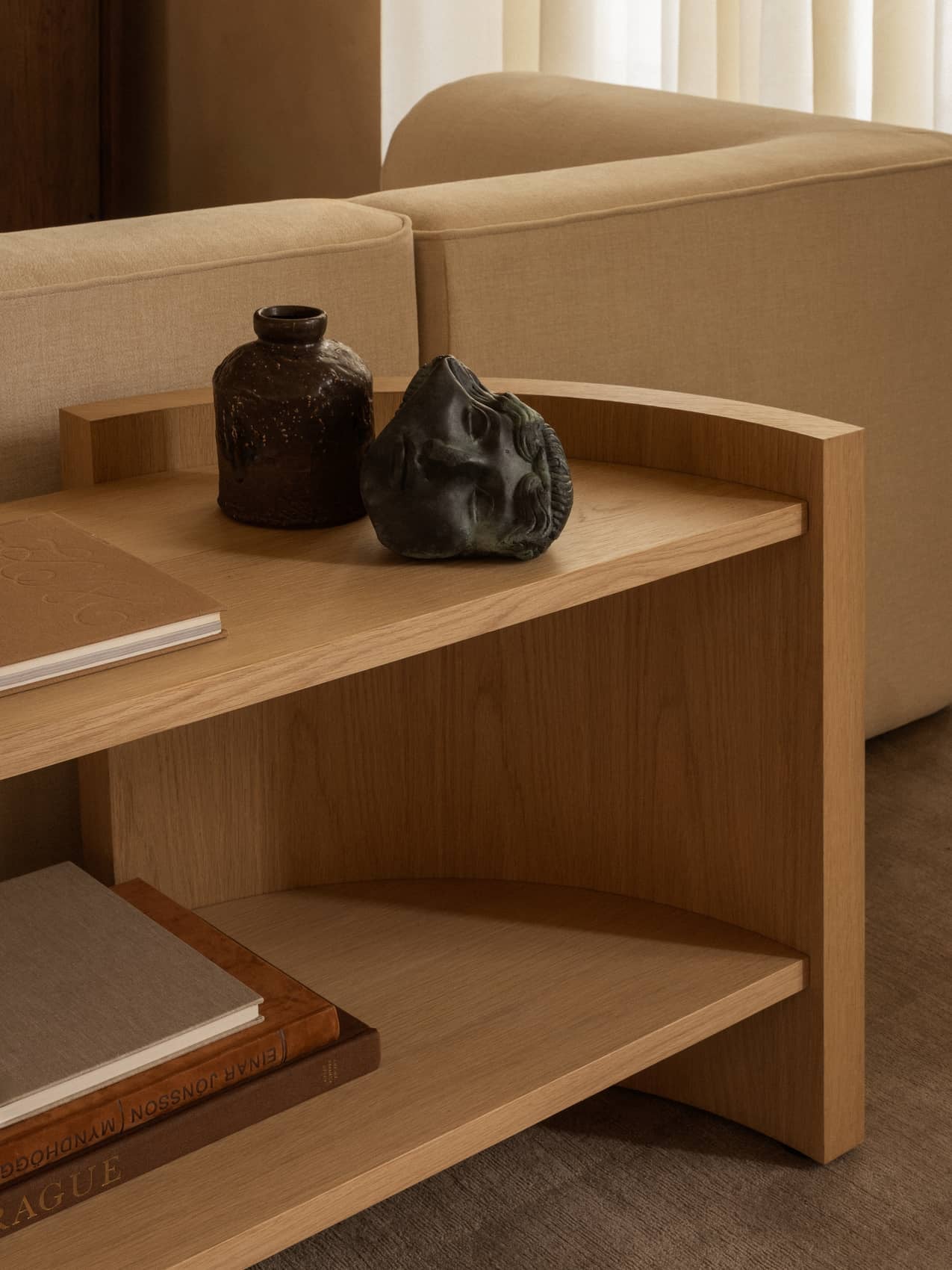 Close-up of the light oak Crescent Shelving H60 styled with vases and books, next to the corner of a beige Eave Modular Sofa.