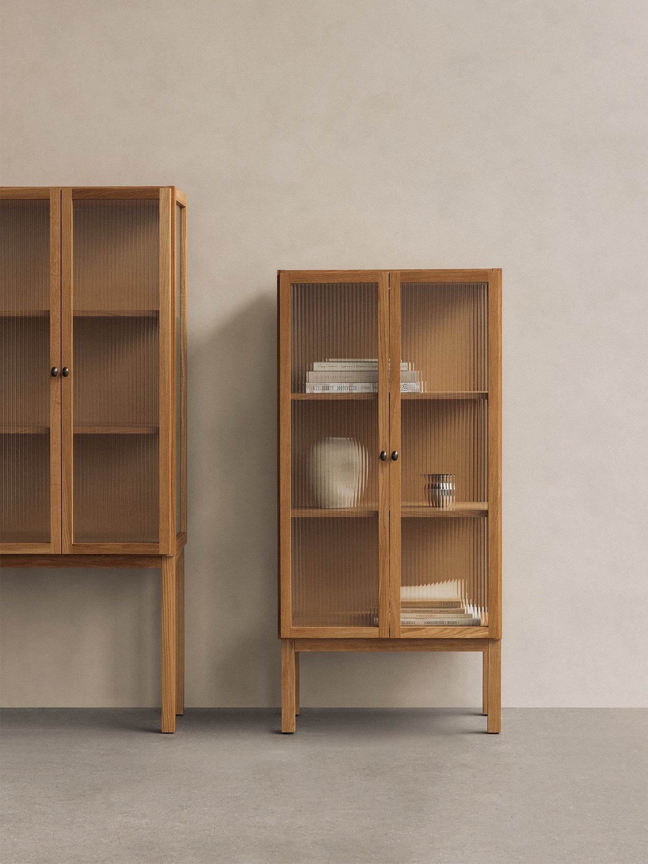 A low oak Curiosity Cabinet with reeded glass doors, filled with books and a vase, beside a taller version in a minimal room.