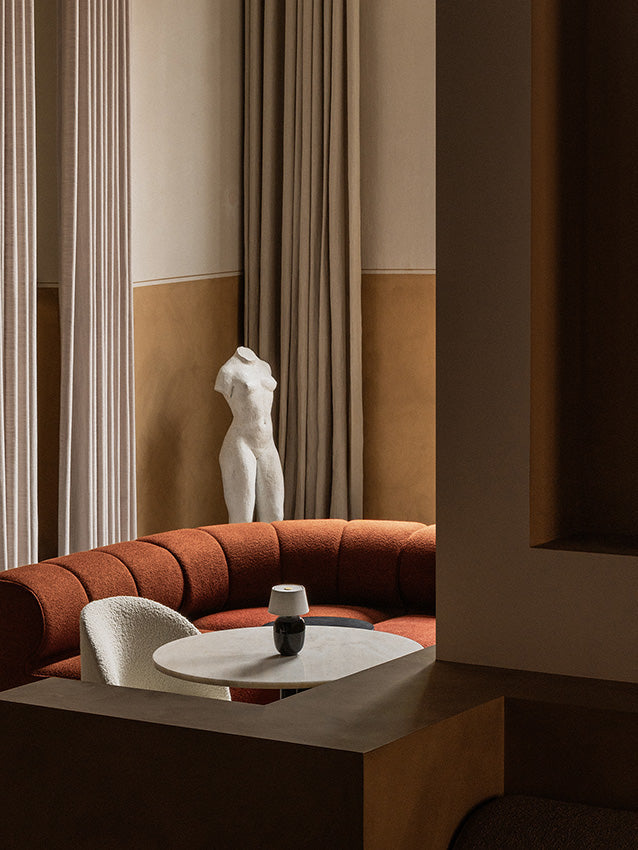 A curved Eave Seamline Modular Sofa in rust orange, styled in a cosy nook with a Nook table, Tearoom chair and Torso lamp.