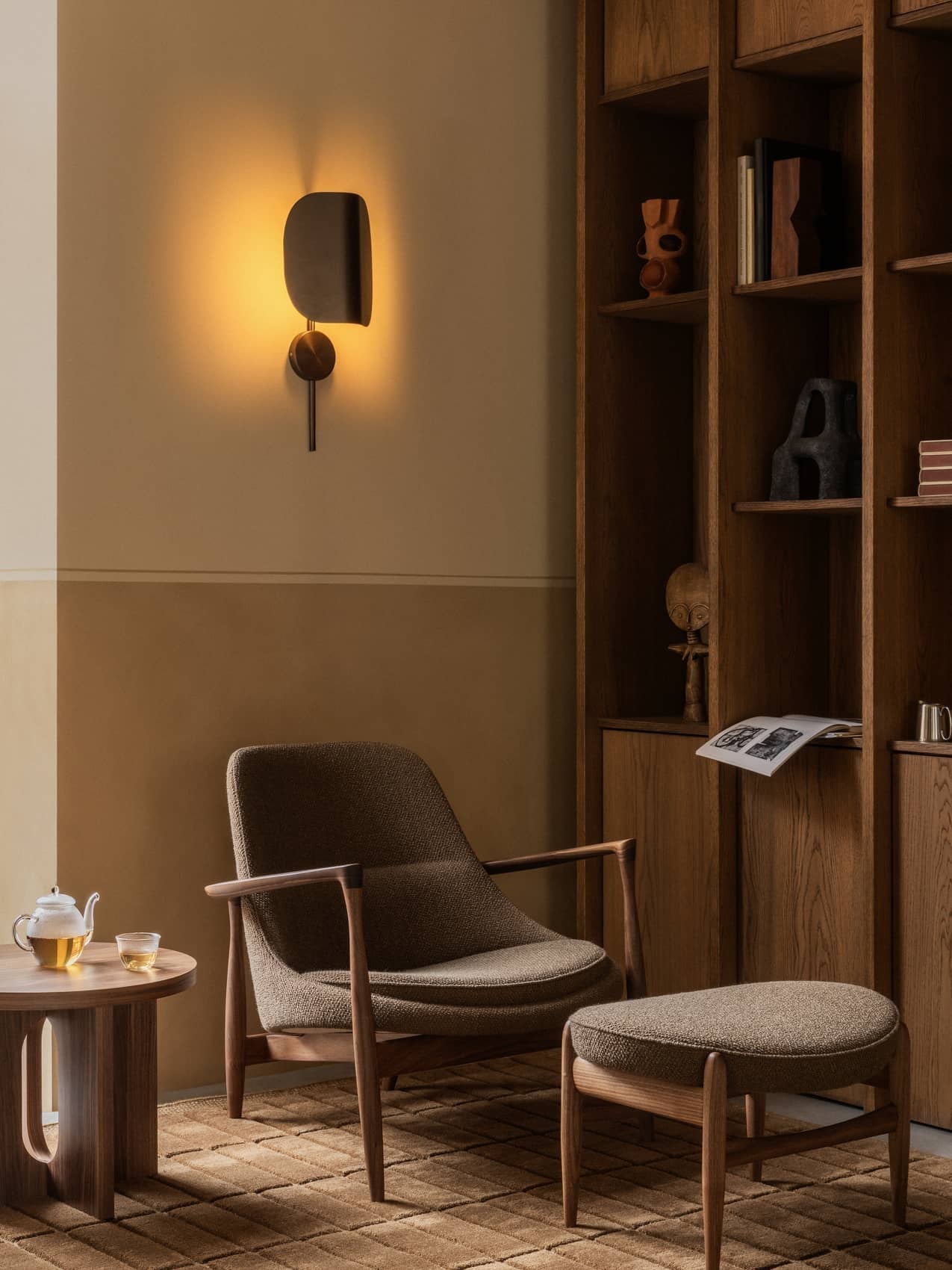 A living room scene with an Elizabeth Lounge Chair, Ottoman, Androgyne Side Table, Tile Rug, and glowing Wing Wall Lamp.