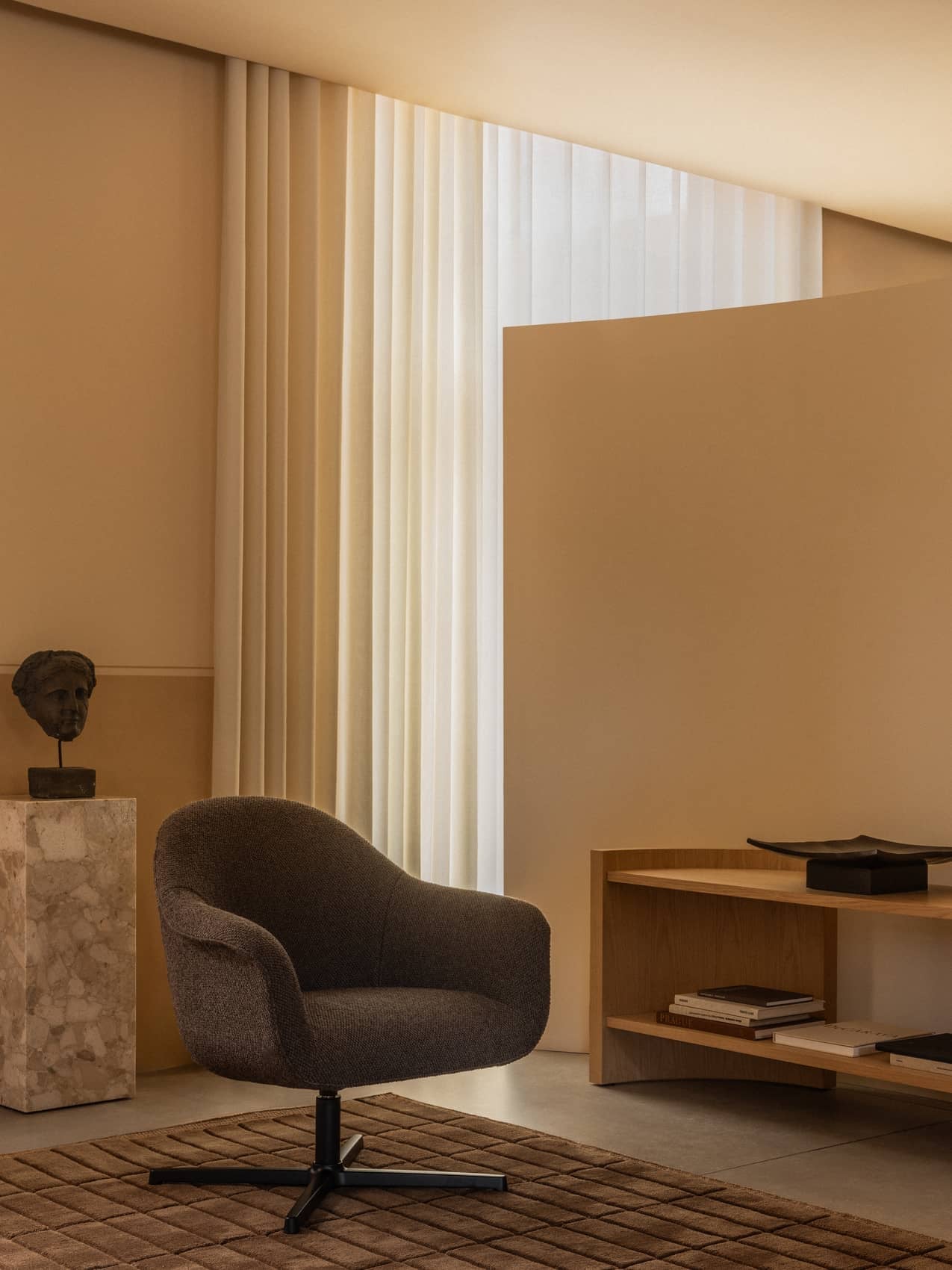 A Harbour Lounge Chair with dark upholstery on a brown Tile Rug in a beige room with Crescent Shelving and a marble plinth.