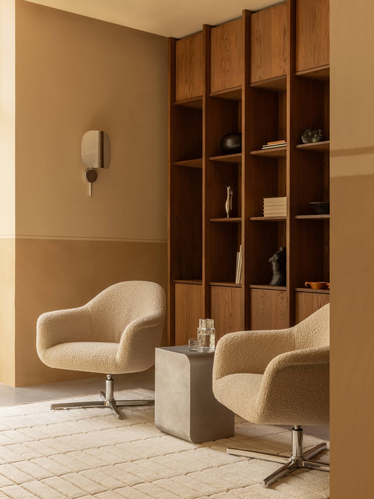 Two bouclé Harbour Lounge Chairs with a Volume Side Table on a Tile Rug. A Wing Wall Lamp hangs by a large wooden bookcase.