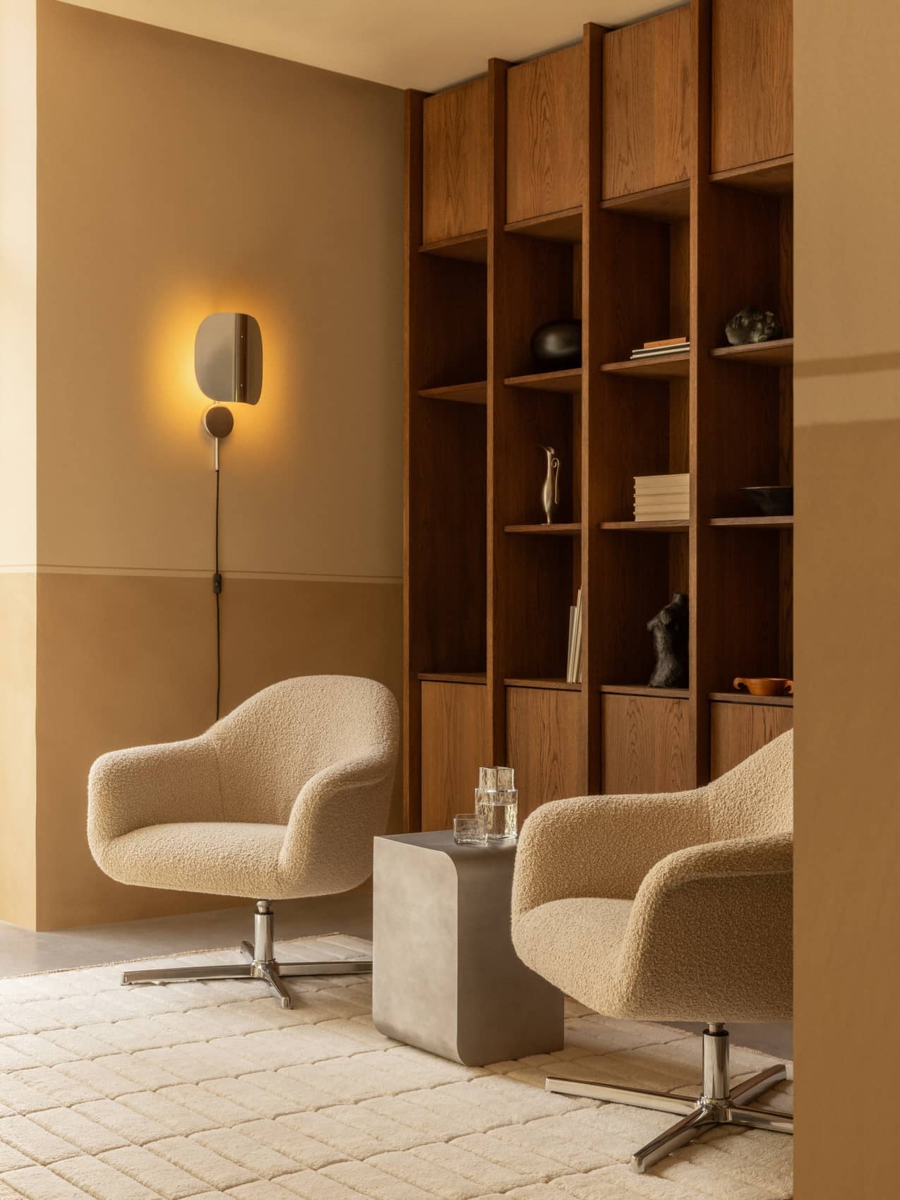 A lounge with a Wing Wall Lamp, two Harbour Lounge Chairs, a Volume Side Table, Tile Rug, and a wooden bookcase.