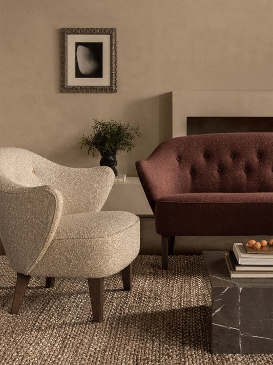 A cream Ingeborg Lounge Chair and a burgundy sofa in a living room with a marble plinth coffee table and a jute rug.