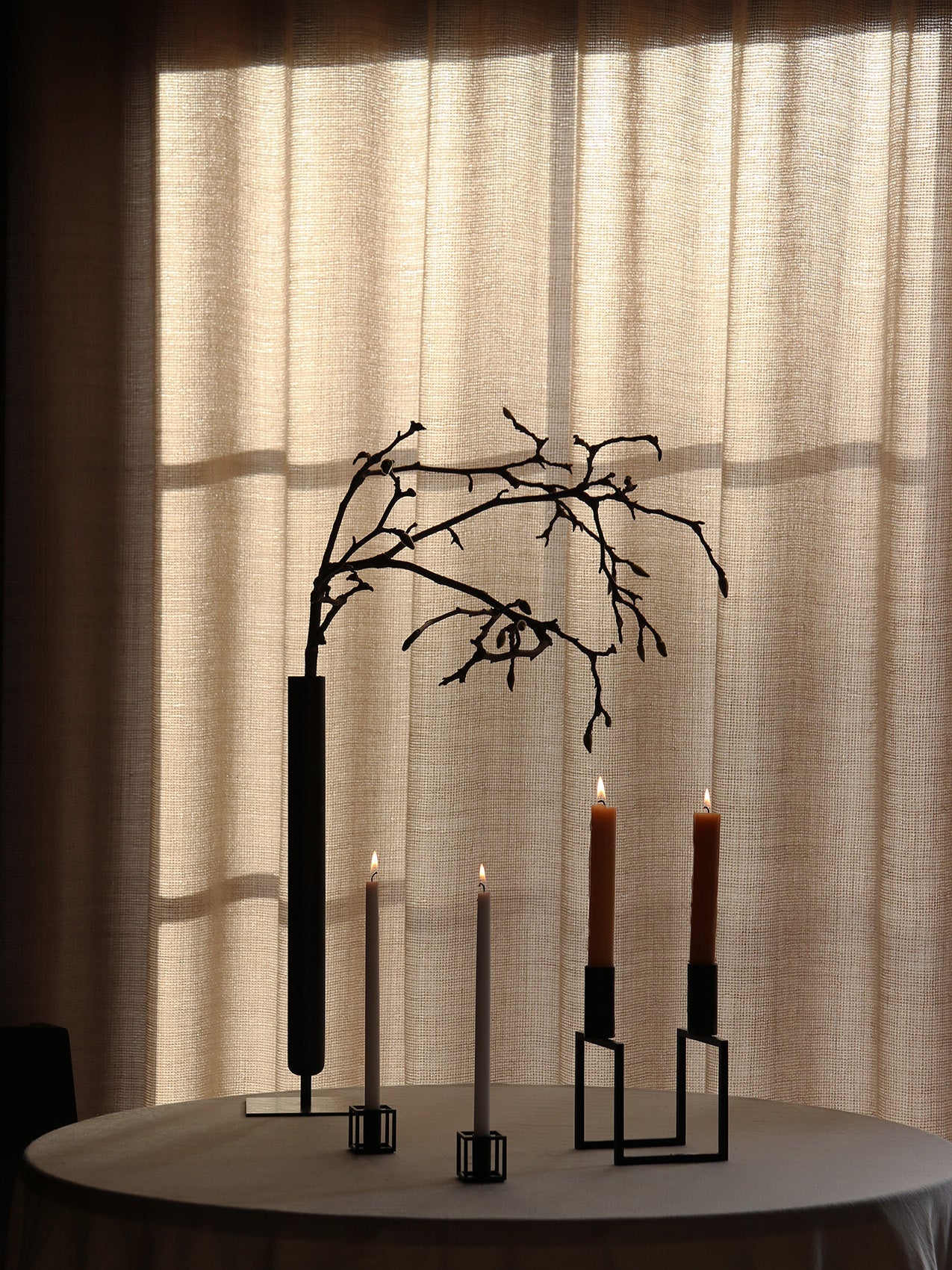 A moody shot of a round table with a tall black Stance Vase, and lit candles in black Kubus Line and Kubus Mikro holders.