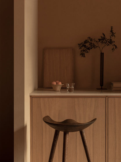 A dark wood ML 42 Counter Stool in a warm, minimalist kitchen. On the counter are a Stance Vase, Triptych Bowl, and glass.