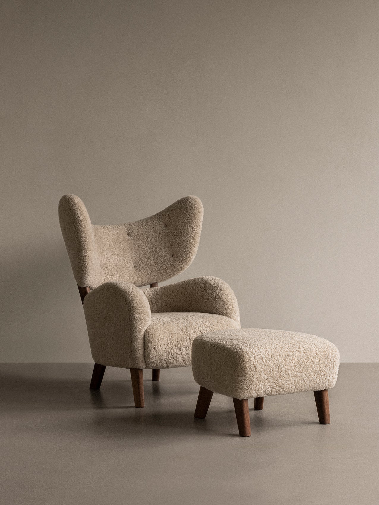 A My Own Chair and matching Ottoman, upholstered in fluffy cream sheepskin with dark wood legs, in a minimally styled room.