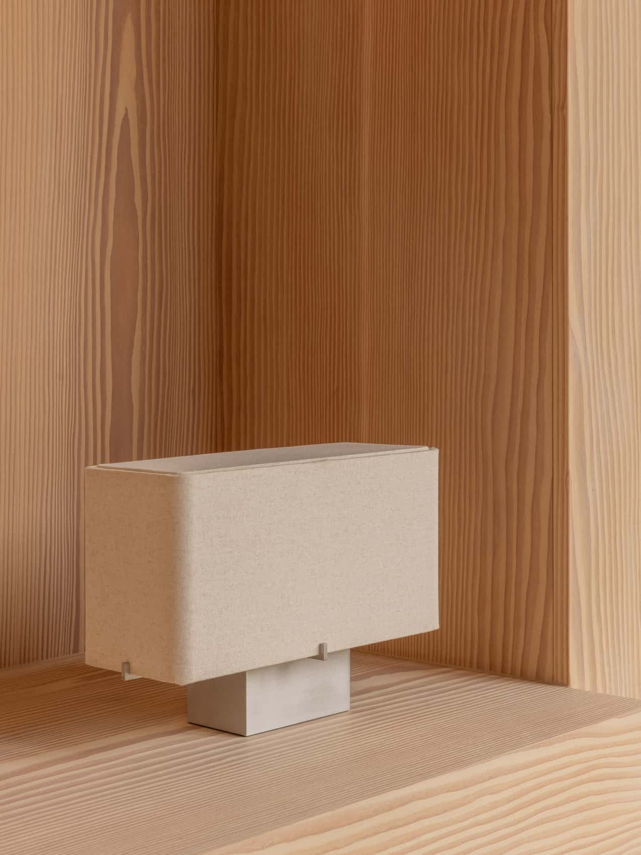 The Nonna Table Lamp, with its rectangular beige textile shade and brushed aluminium base, resting in a light wood alcove.