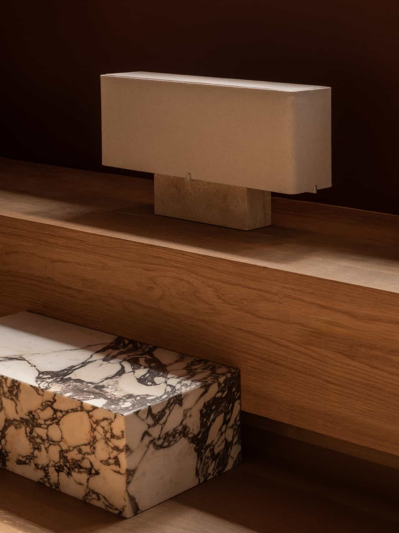 The Nonna Table Lamp in travertine with its rectangular sand-coloured shade, on a wooden shelf with a marble block below.