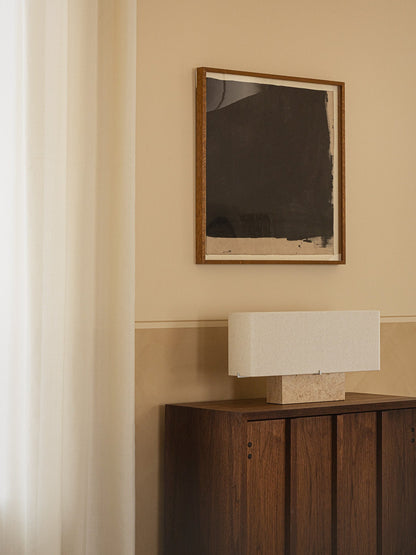 Nonna Table Lamp, travertine base and rectangular shade, sits on a dark wood sideboard beneath an abstract painting.