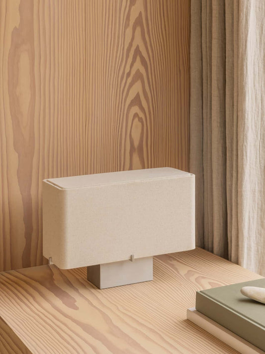 Nonna Table Lamp with a rectangular sand-coloured shade on a light wood desk against a matching wood-panelled wall.