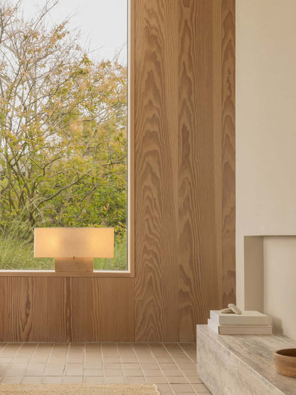 The wide Nonna Table Lamp in travertine sits lit on a wooden windowsill, casting a warm glow in a room with wood panelling.