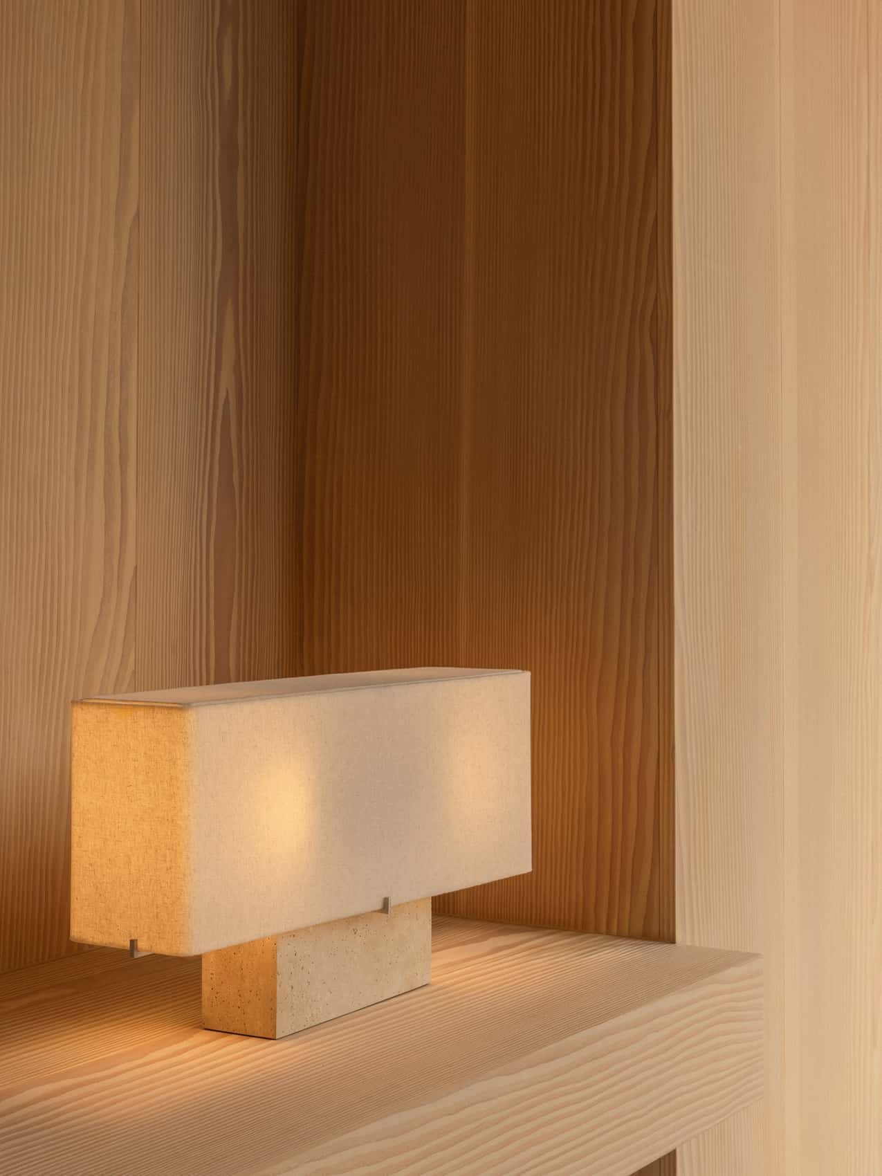 The wide Nonna Table Lamp in travertine sits on a light wood shelf, its lit fabric shade casting a warm glow in a corner.