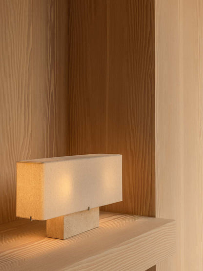 The wide Nonna Table Lamp in travertine sits on a light wood shelf, its lit fabric shade casting a warm glow in a corner.