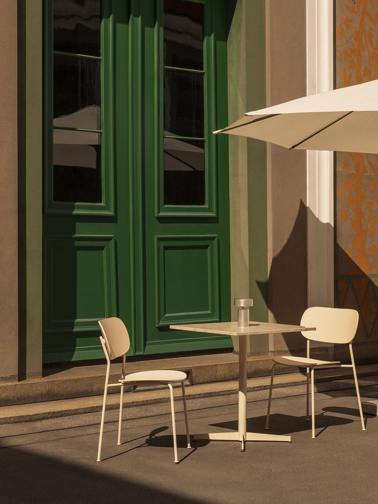 A sunlit patio with an ivory Nook Dining Table, two Co Dining Chairs, and a Column Portable lamp before a large green door.