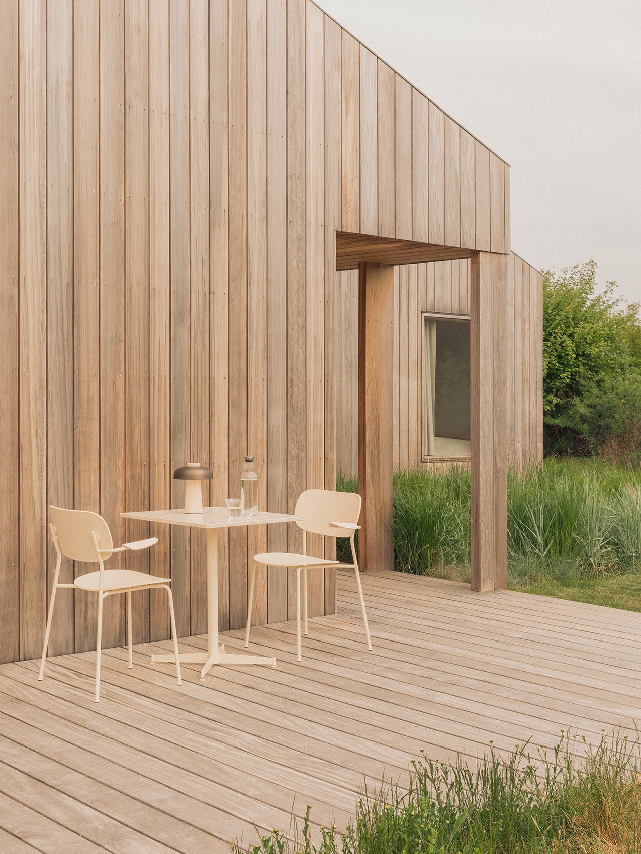 An ivory Nook Dining Table and two Co Dining Chairs on a wooden deck with a Reverse Portable Lamp and Bottle Carafe on top.
