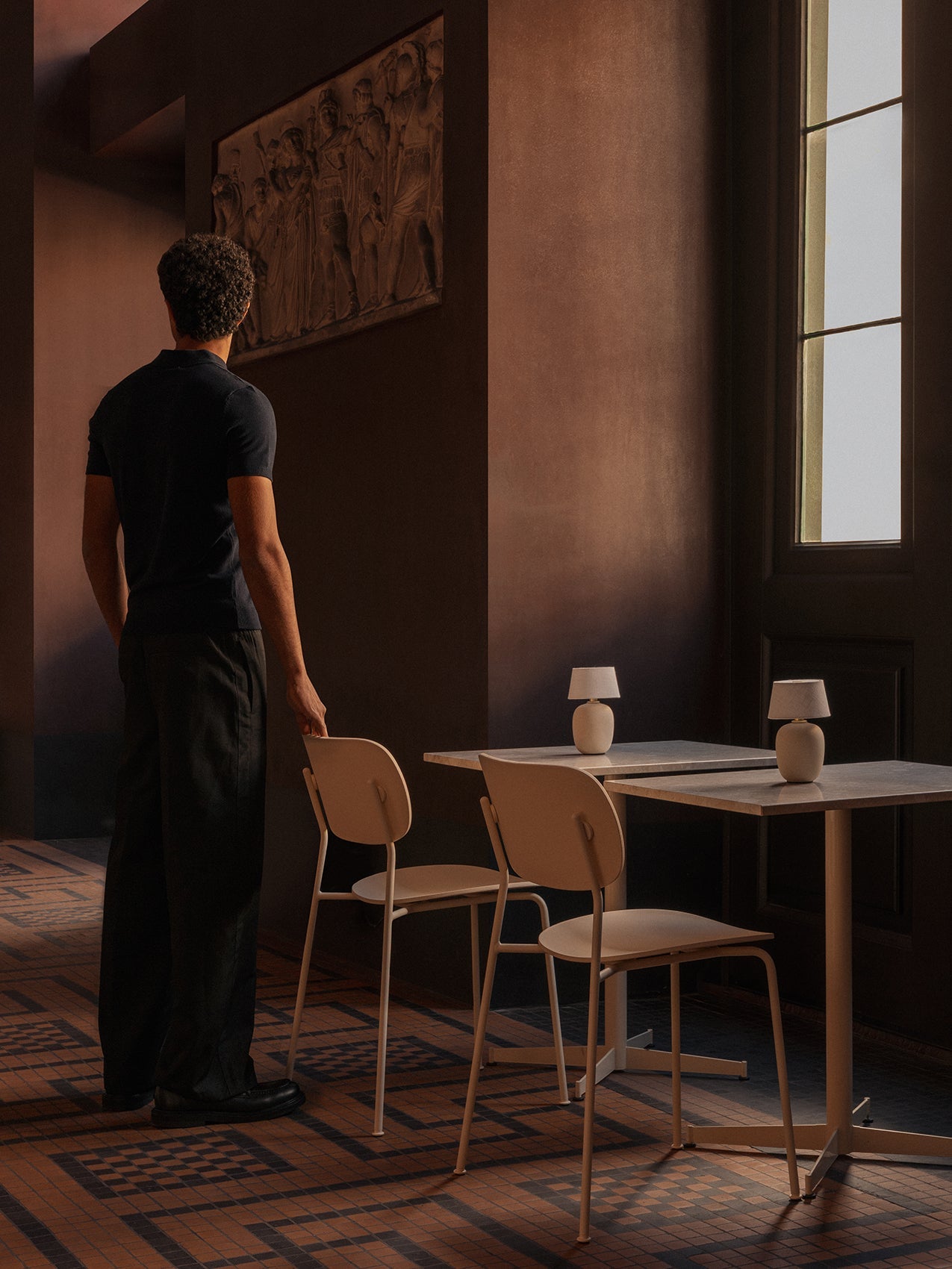 A man stands in a dark room by two ivory Nook Dining Tables and Co Dining Chairs, with Torso Portable lamps on the tables.