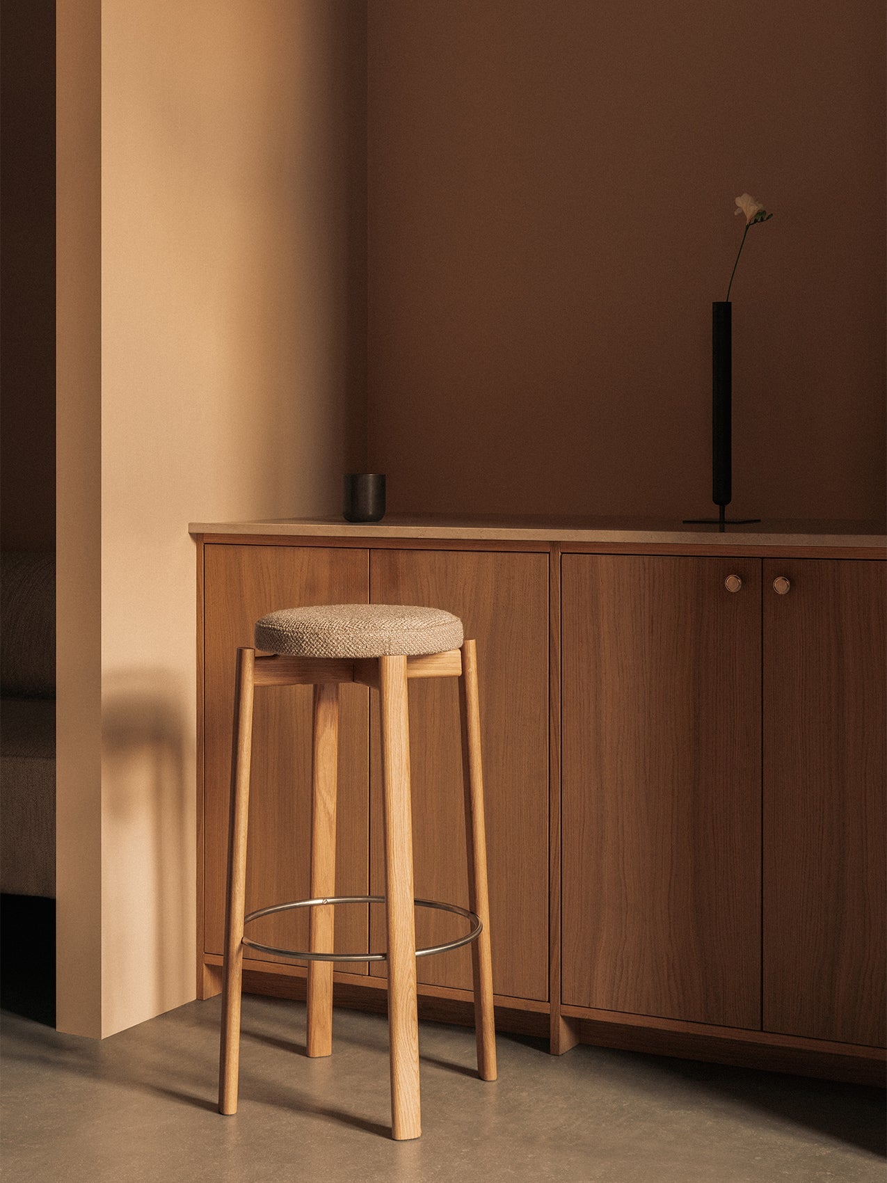 An oak Passage Bar Stool with an upholstered seat stands in a warm-toned corner next to a wooden cabinet with a vase on it.