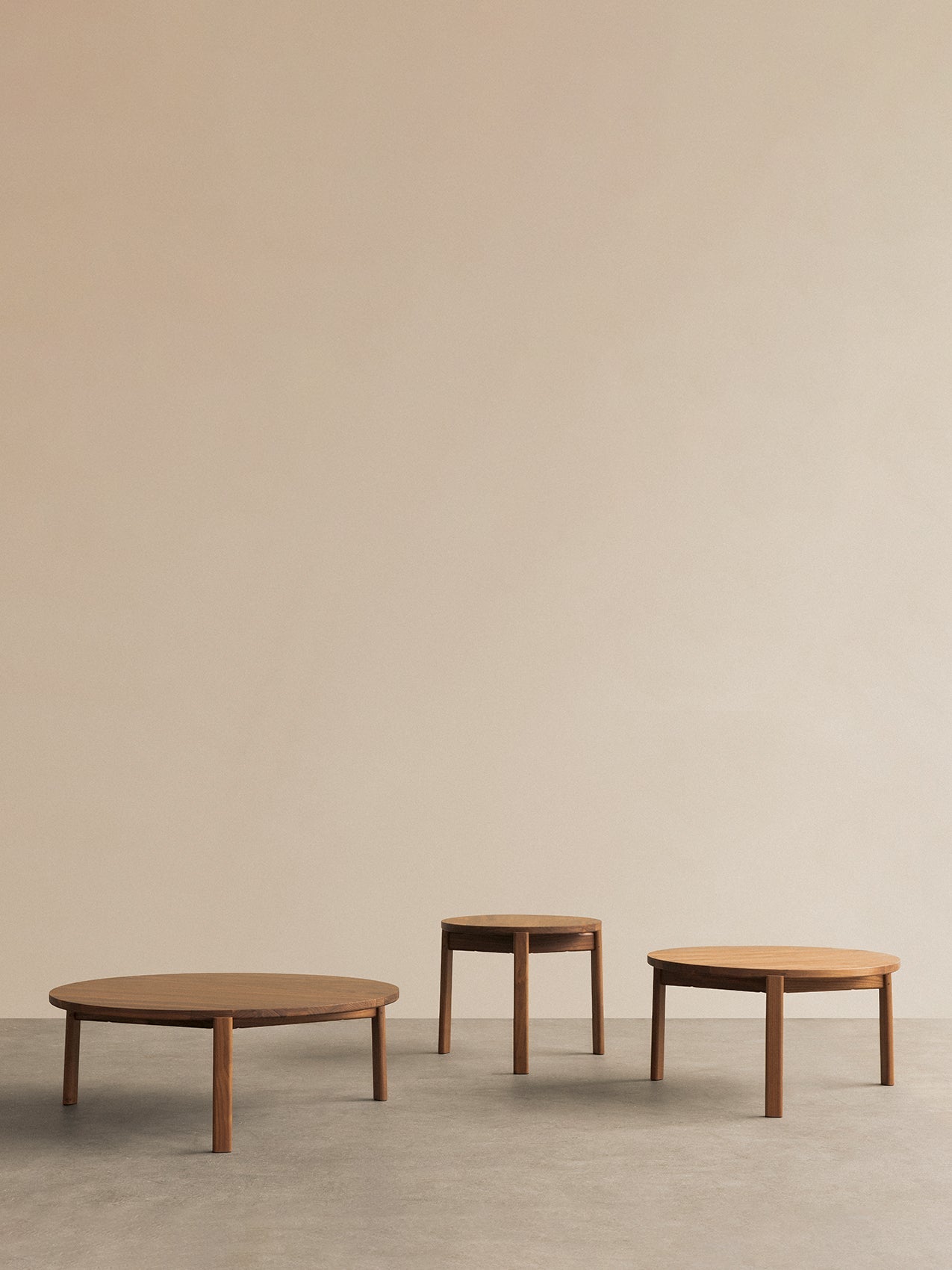 A set of three round, wooden Passage Lounge Tables in varying sizes, set against a plain beige wall on a grey floor.