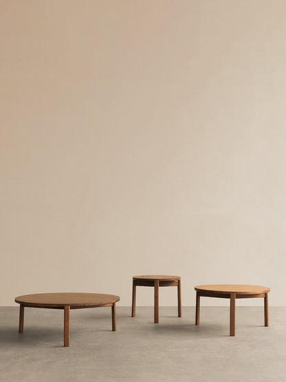A set of three round, wooden Passage Lounge Tables in varying sizes, set against a plain beige wall on a grey floor.