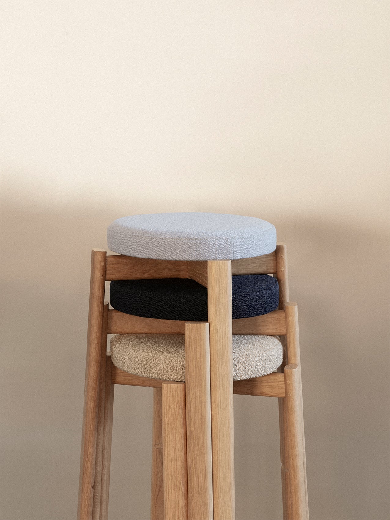 A close-up of three stackable Passage Stools in natural oak, with upholstered seats in light grey, dark blue, and beige.