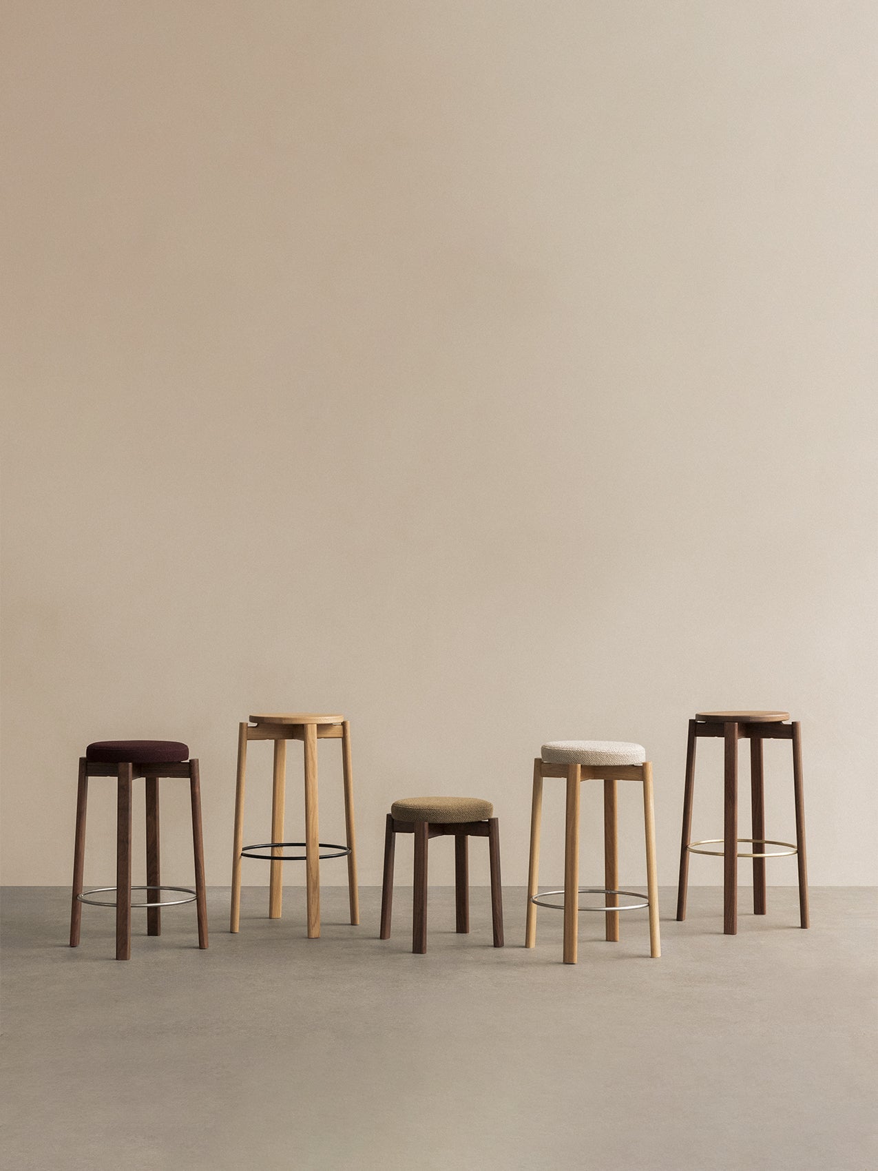 Five Passage Counter Stools in various heights, wood finishes like oak & walnut, and upholstered seats against a beige wall.