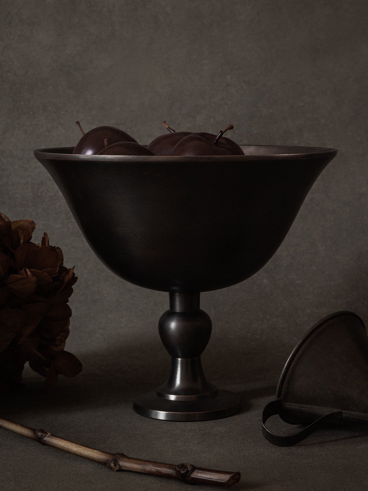 A still life image of the dark bronzed Pavot Bowl on a pedestal, filled with dark plums, set against a dark grey background.