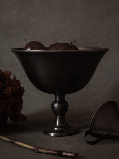 A still life image of the dark bronzed Pavot Bowl on a pedestal, filled with dark plums, set against a dark grey background.