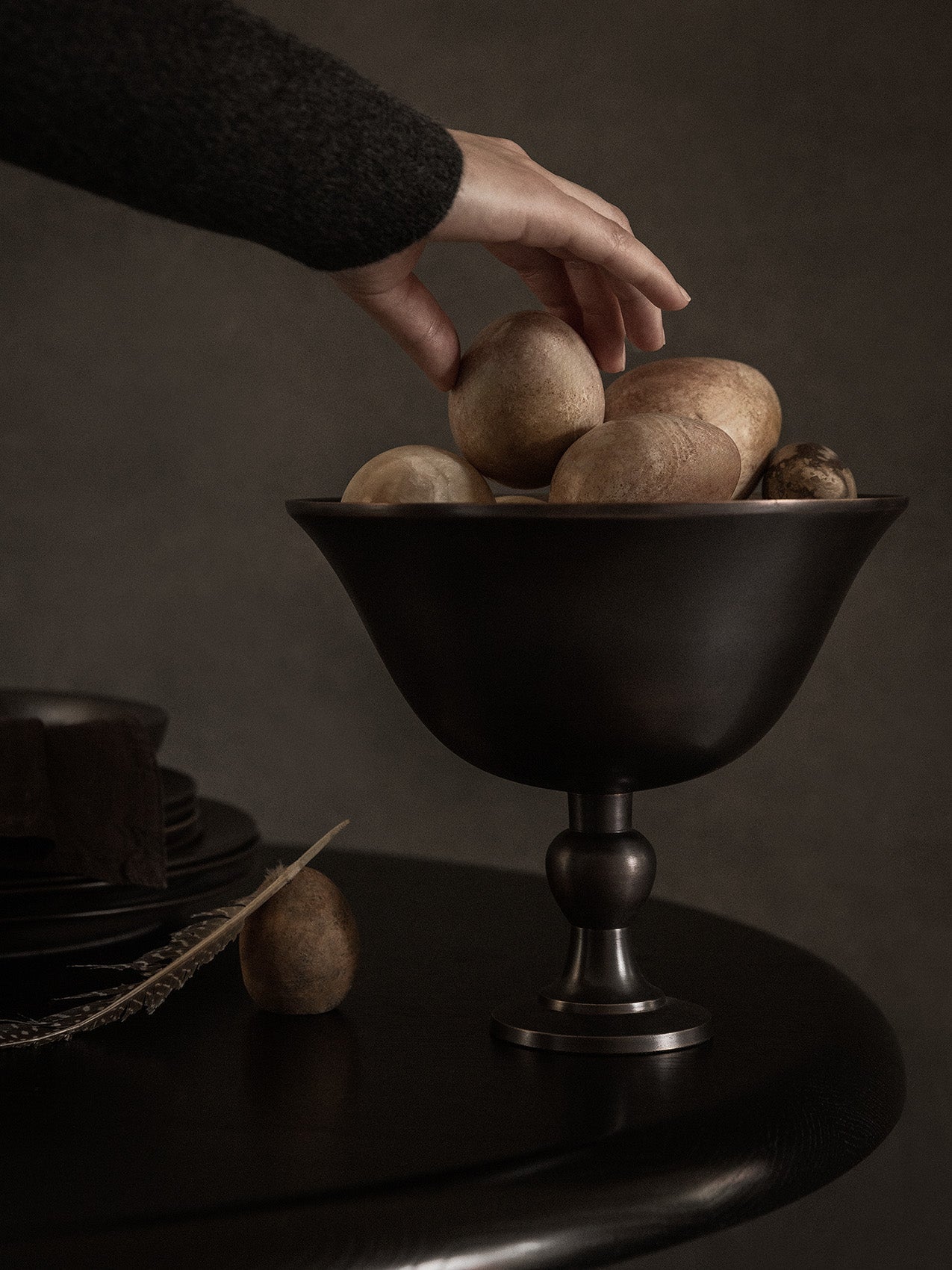 A hand in a dark sweater reaches for a stone inside the dark bronze Pavot Bowl, which sits on a table in a moody setting.
