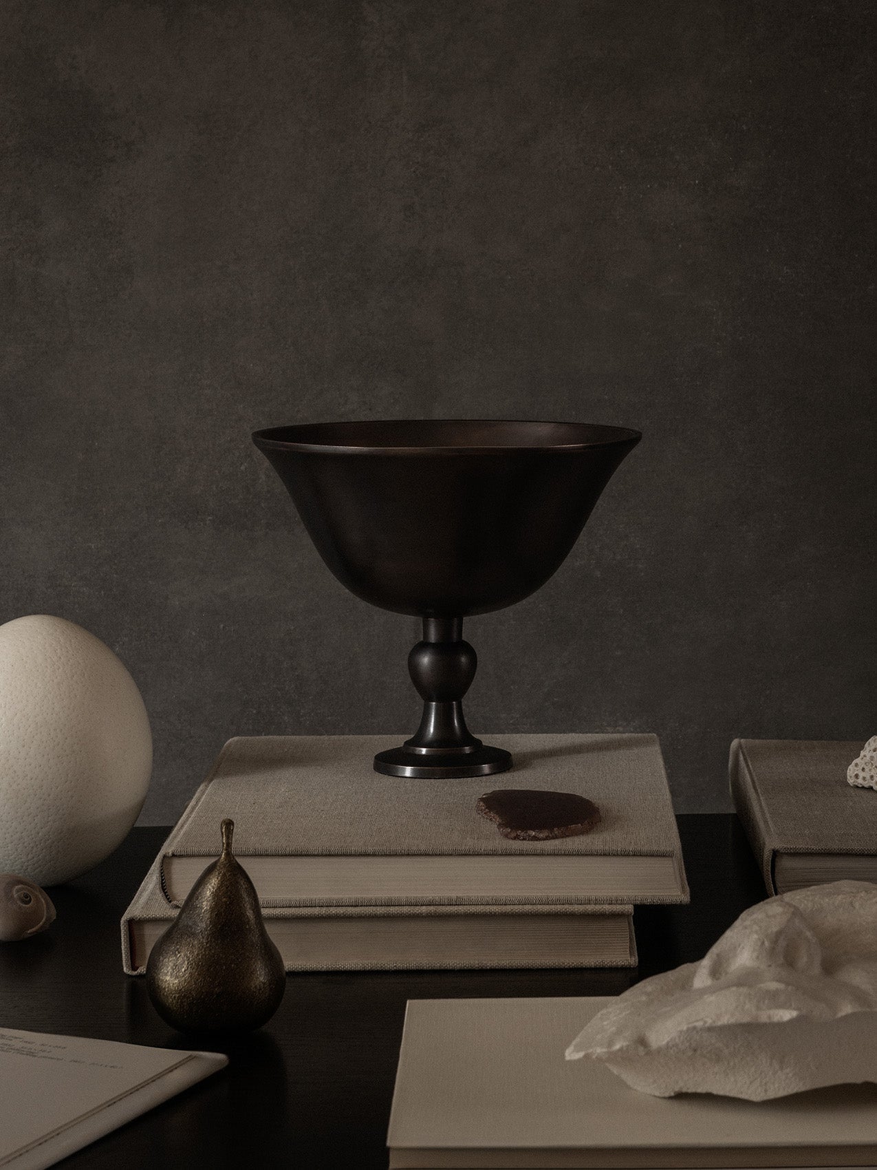 The Audo Pavot Bowl in a dark finish, styled on a stack of books in a moody still life with a bronze pear and an ostrich egg.
