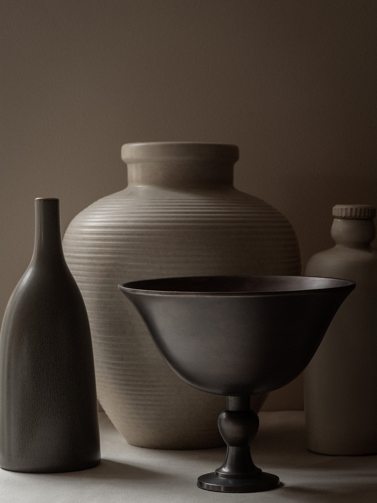 The dark bronze Pavot Bowl on a pedestal in a still life with a slender dark vase and two larger, textured beige vases.