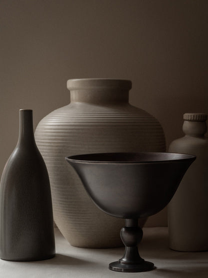 The dark bronze Pavot Bowl on a pedestal in a still life with a slender dark vase and two larger, textured beige vases.