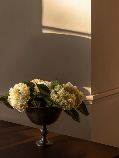 The Audo Pavot Bowl, with white and green hydrangeas, on a wooden table. Warm light casts a shadow on the wall.